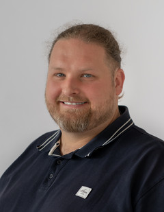 Porträt eines Mannes mit kurzem Haar und Bart, der ein dunkles Poloshirt mit kleinem Logo trägt, vor hellem Hintergrund