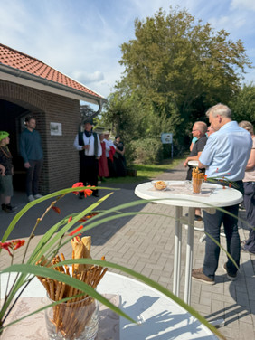 Menschen stehen an Stehtischen im Freien neben einem Gebäude mit rotem Ziegeldach, im Hintergrund Bäume und eine Gruppe in Trachtenkleidung