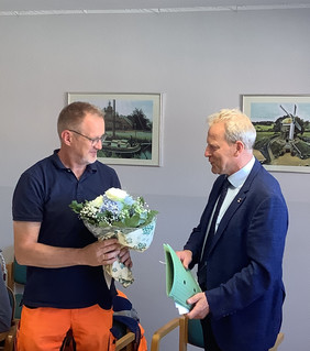 Zwei Männer stehen in einem Raum, einer hält einen Blumenstrauß.