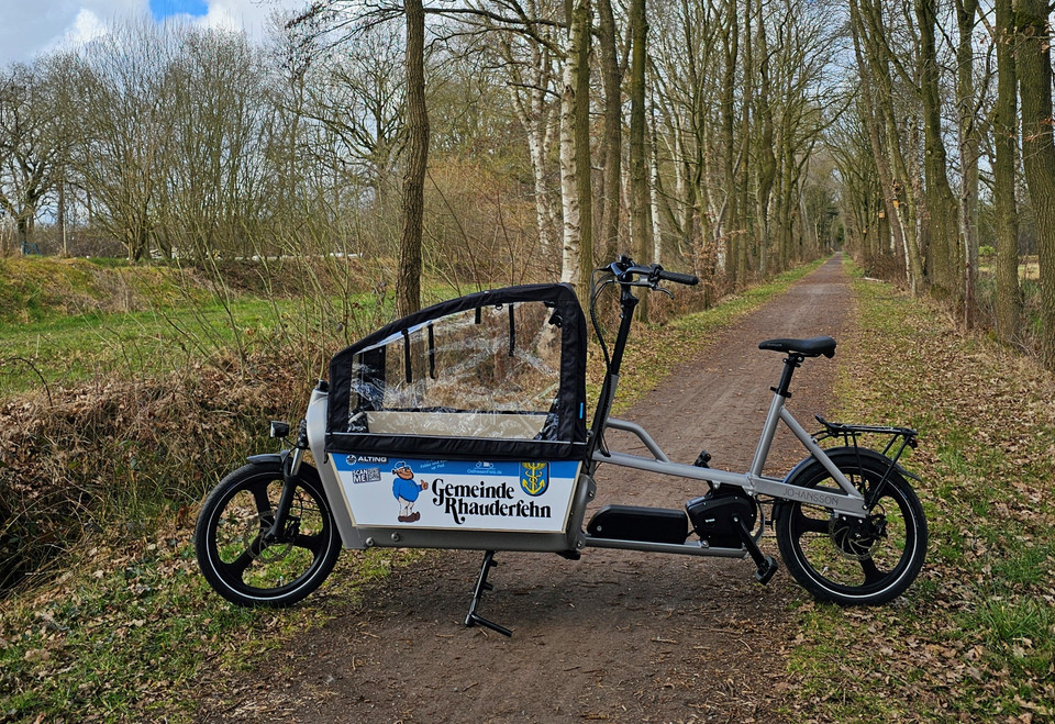 Ein Lastenfahrrad steht quer auf einem Wanderweg. Es ist mit dem Wappen der Gemeinde Rhauderfehn und dem Gemeindemaskottchen Fokko bedruckt.