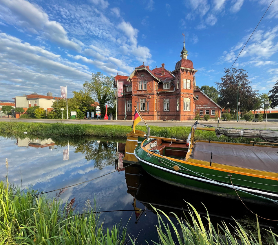 Tjalk mit deutscher Flagge am Ufer eines Kanals, im Hintergrund ein rotes Backsteingebäude mit Turm und Schildern, blauer Himmel mit Wolken.