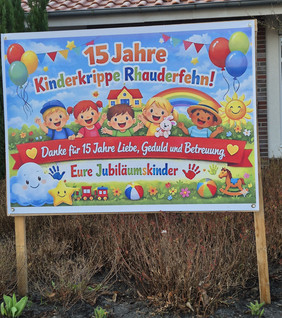 Buntes Schild mit fünf lachenden Kindern vor Haus und Regenbogen, Luftballons, Sonne, Wolke, Stern, Spielzeug und Text zum 15-jährigen Jubiläum der Kinderkrippe Rhauderfehn