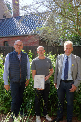 Drei Personen stehen im Garten vor einem Haus, umgeben von Pflanzen und Bäumen