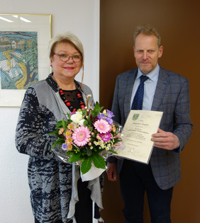 Zwei Personen stehen nebeneinander, eine hält einen Blumenstrauß, die andere eine Urkunde