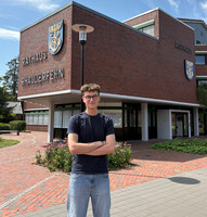 Junger Mann mit Brille, schwarzem T-Shirt und blauer Jeans steht mit verschränkten Armen vor dem roten Backsteingebäude des Rathauses Rhauderfehn unter blauem Himmel