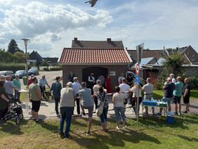Menschen versammeln sich im Freien vor einem kleinen Gebäude mit rotem Ziegeldach an einem sonnigen Tag