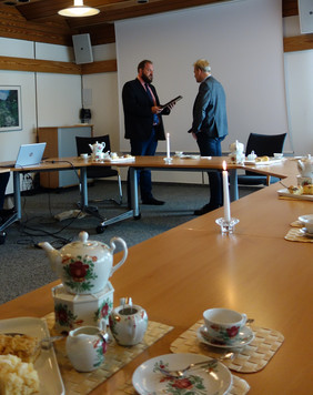 Zwei Männer stehen sich in einem Raum mit Tischen und Stühlen gegenüber, auf einem Tisch im Vordergrund stehen Kaffeetassen und ein Blumenstrauß