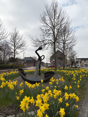Ankerskulptur in einem Beet mit blühenden Narzissen vor kahlen Bäumen und einem Haus im Hintergrund