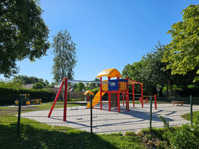 Spielplatz mit Klettergerüst und Rutsche, umgeben von grünem Zaun und Bäumen bei klarem Himmel