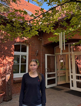 Person mit langem Haar und schwarzem Langarmshirt steht vor dem Eingang eines Backsteingebäudes mit offenem Glastür und Fenstern