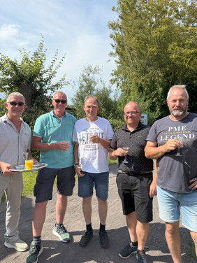Fünf Männer stehen draußen auf einem Weg, umgeben von Bäumen, tragen Sommerkleidung und halten Getränke in der Hand