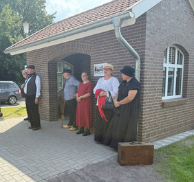 Fünf Personen in historischer Kleidung stehen vor einem Backsteingebäude mit rotem Ziegeldach und einem Schild neben der Tür
