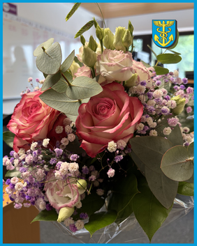 Nahaufnahme eines Blumenstraußes mit Rosen, Schleierkraut und Eukalyptusblättern, im Hintergrund unscharf ein Wappen mit blauem Schild und gelbem Symbol