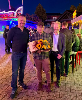 Drei Männer stehen nebeneinander, der mittlere hält einen Blumenstrauß und eine Urkunde, alle tragen Freizeitkleidung 