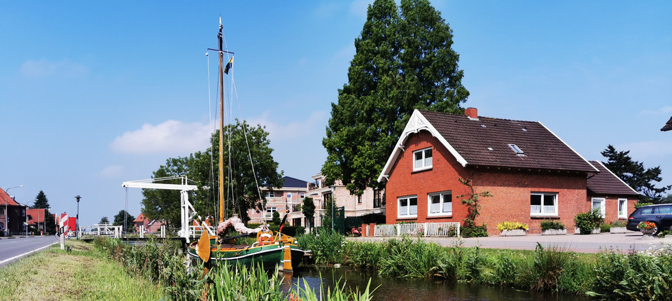 Grüne Tjalk mit Mast am Ufer eines Kanals, daneben rotes Backsteinhaus mit Schornstein und großem Baum bei blauem Himmel.