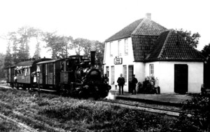 Dampflokomotive mit mehreren Waggons an einem kleinen Bahnhof mit zwei Personen vor dem Gebäude mit Schildern.