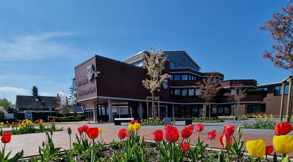 Backsteingebäude der Gemeinde Rhauderfehn mit Schriftzug und Wappen, davor bunte Tulpen und gepflasterter Platz unter blauem Himmel.