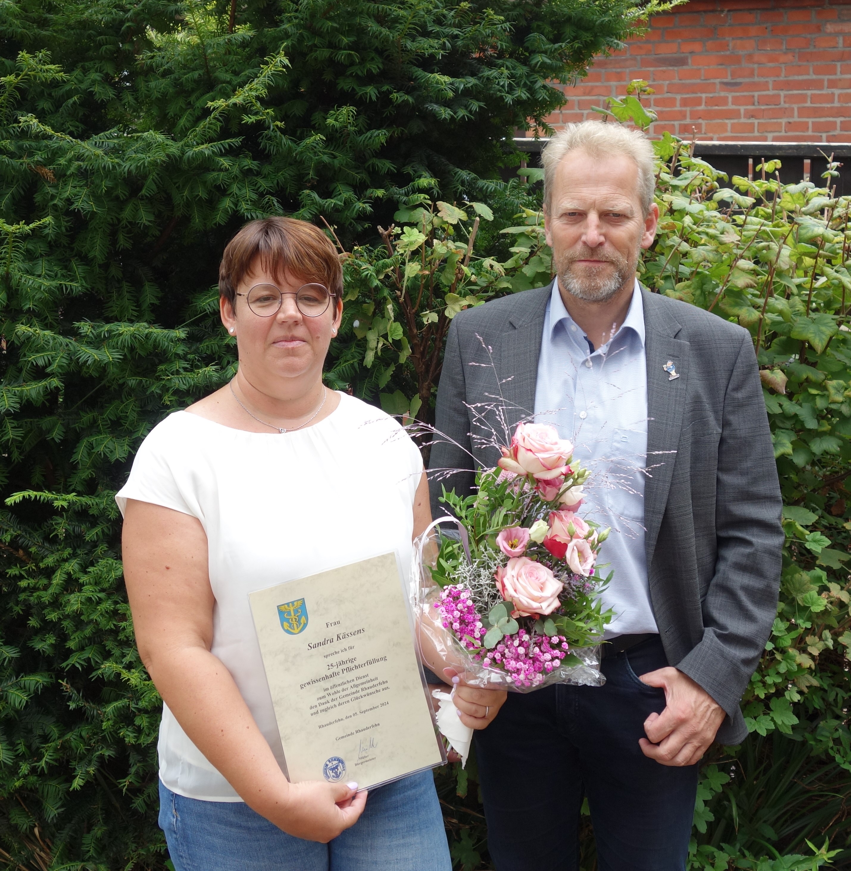 Zwei Personen stehen vor einem grünen Busch, die Frau hält eine Urkunde, der Mann einen Blumenstrauß