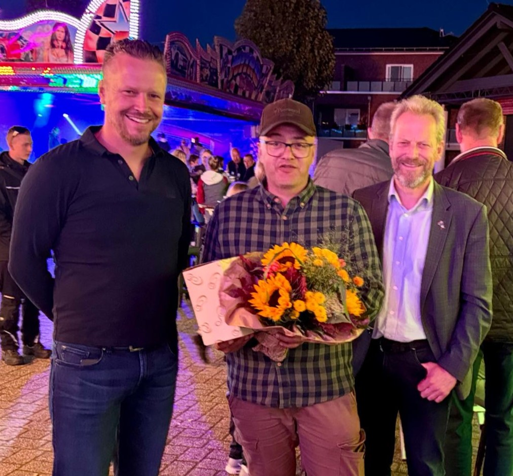 Drei Männer stehen nebeneinander, der mittlere hält einen Blumenstrauß und eine Urkunde, alle tragen Freizeitkleidung 