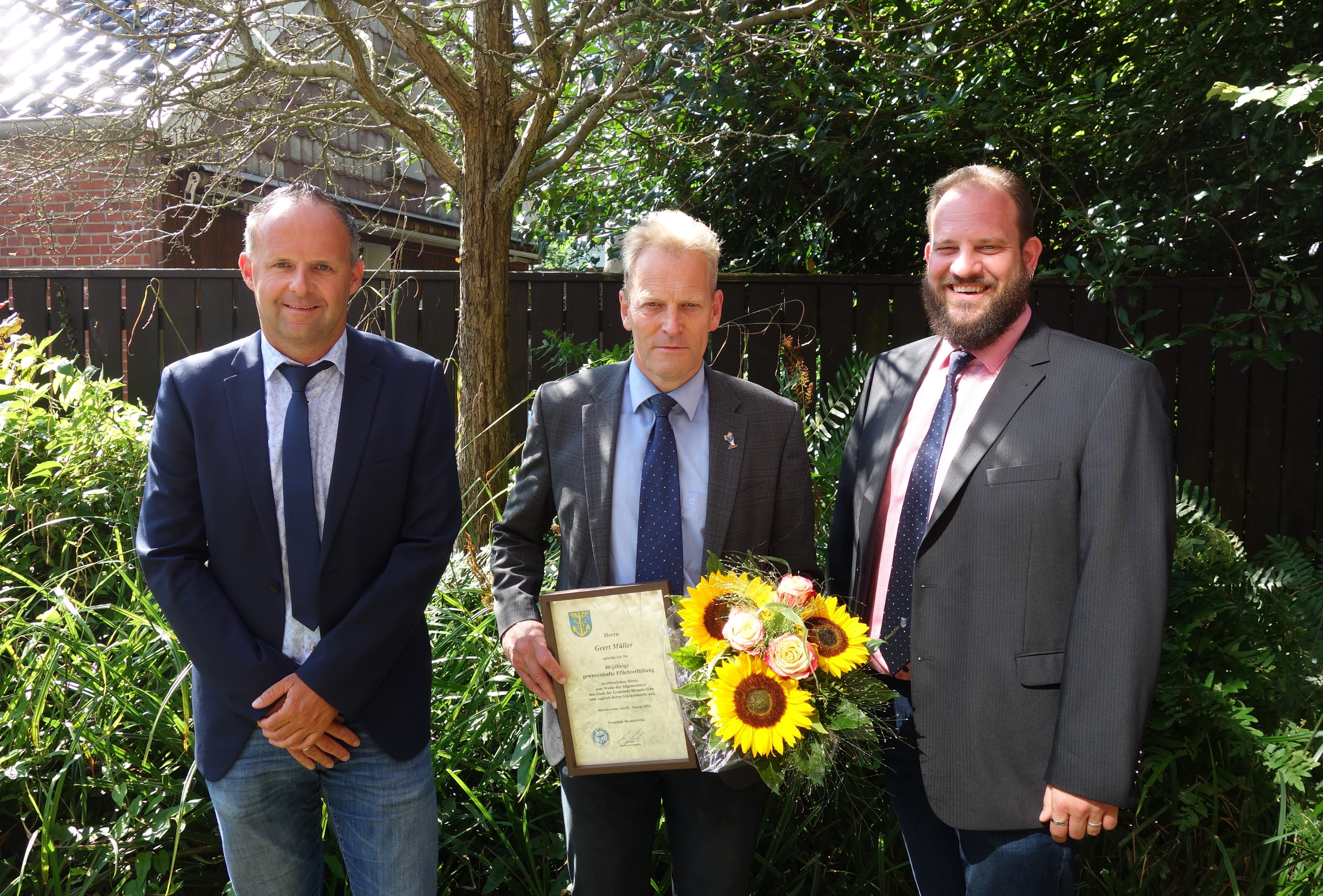 Drei Männer in Anzügen stehen vor einem grünen Busch, der mittlere hält eine Urkunde und einen Blumenstrauß mit Sonnenblumen