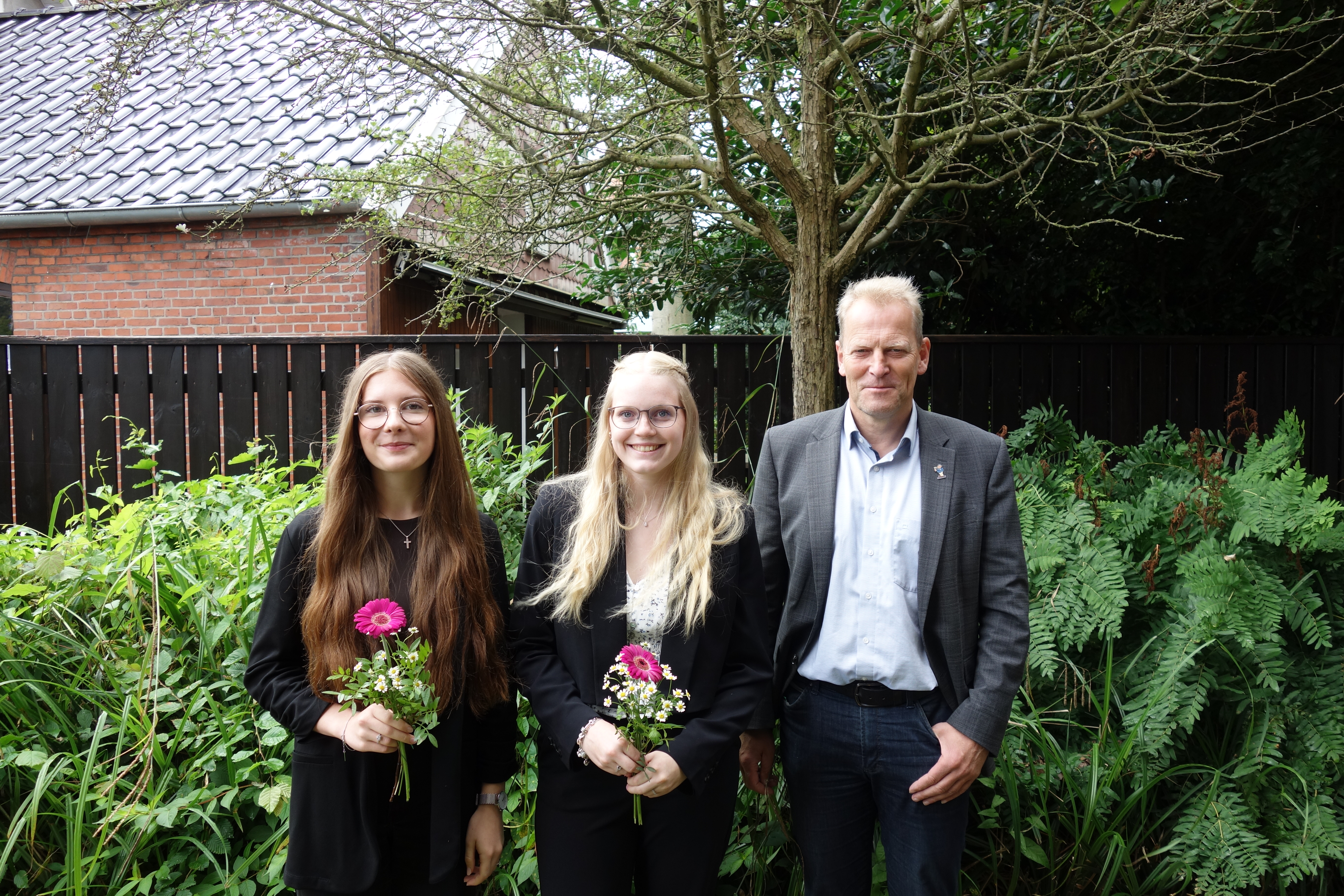 Drei Personen stehen nebeneinander im Garten vor einem Zaun und einem Baum, zwei Frauen halten jeweils eine Gerbera in der Hand.