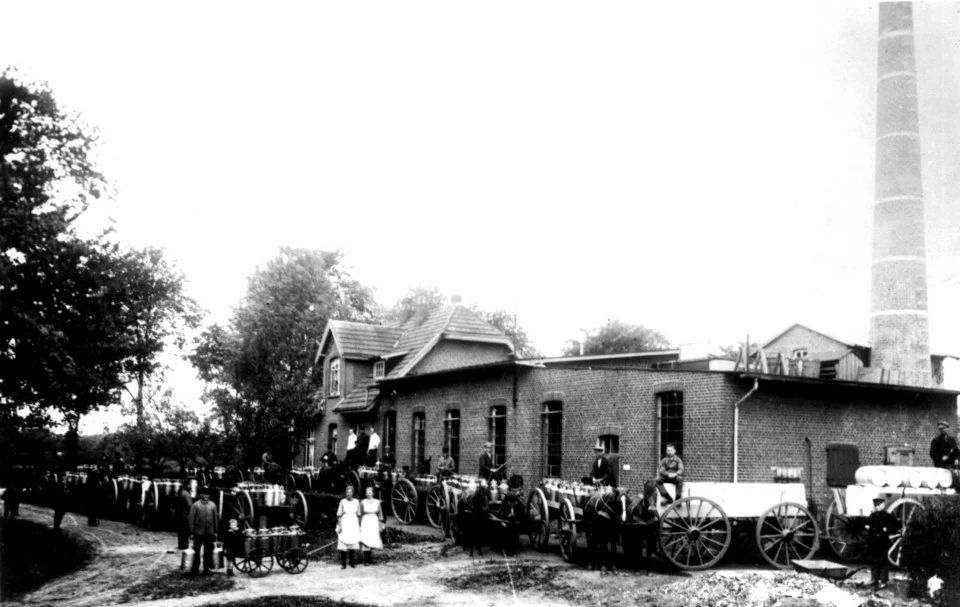 Historisches Foto einer Molkerei mit mehreren Pferdekutschen und Personen vor einem Backsteingebäude mit Schornstein.