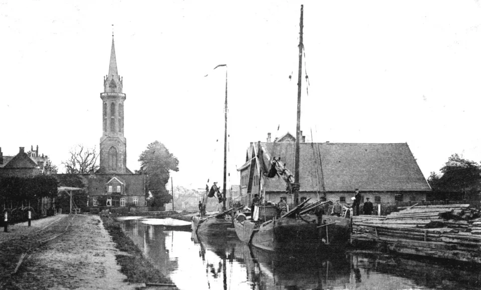 Schwarz-weiß-Foto eines Kanals mit einem Segelschiff am Ufer, daneben Holzstapel und ein hoher Kirchturm im Hintergrund.